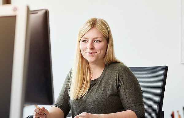 Eine junge, blonde Frau sitzt am Schreibtisch und blickt auf den Bildschirm, während sie auf einem Block etwas notiert.