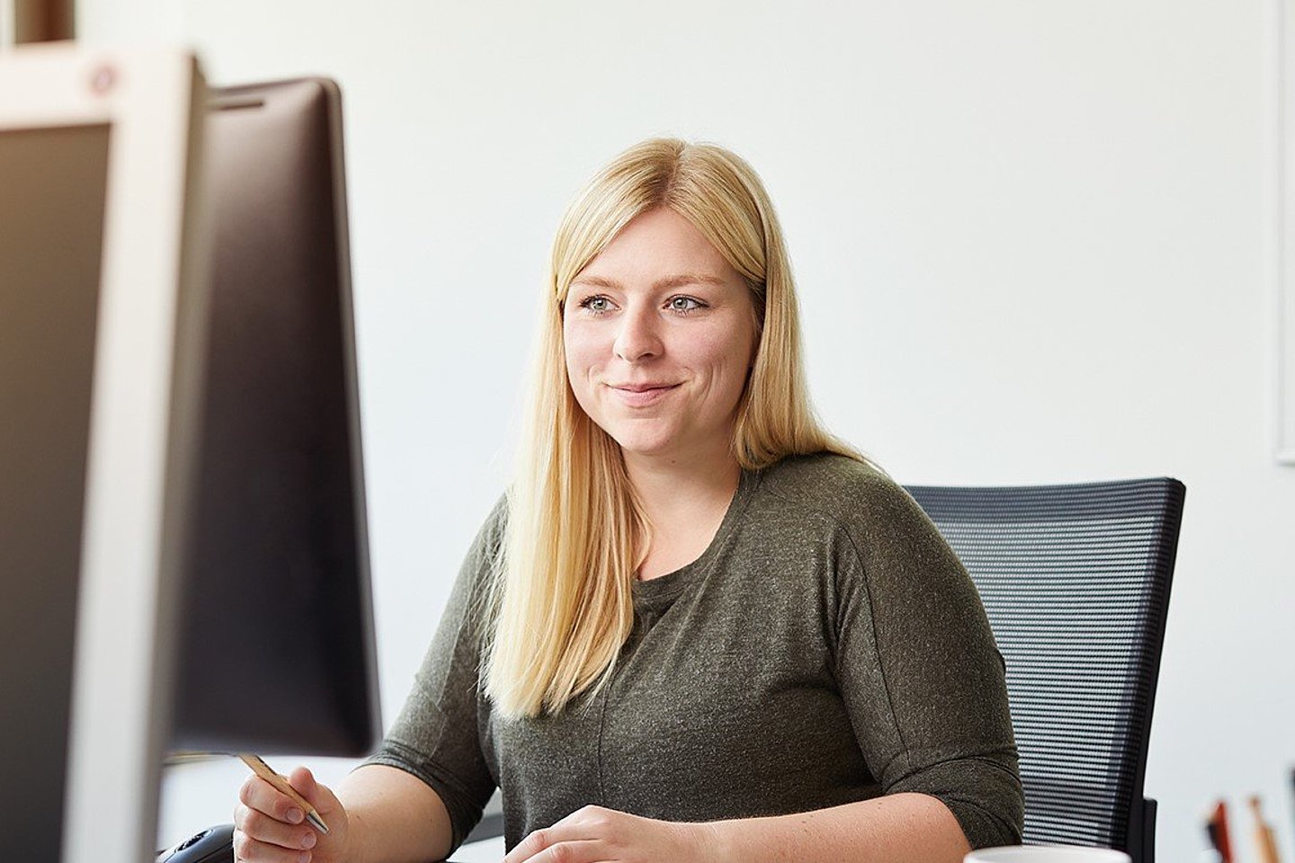 Eine junge, blonde Frau sitzt am Schreibtisch und blickt auf den Bildschirm, während sie auf einem Block etwas notiert.