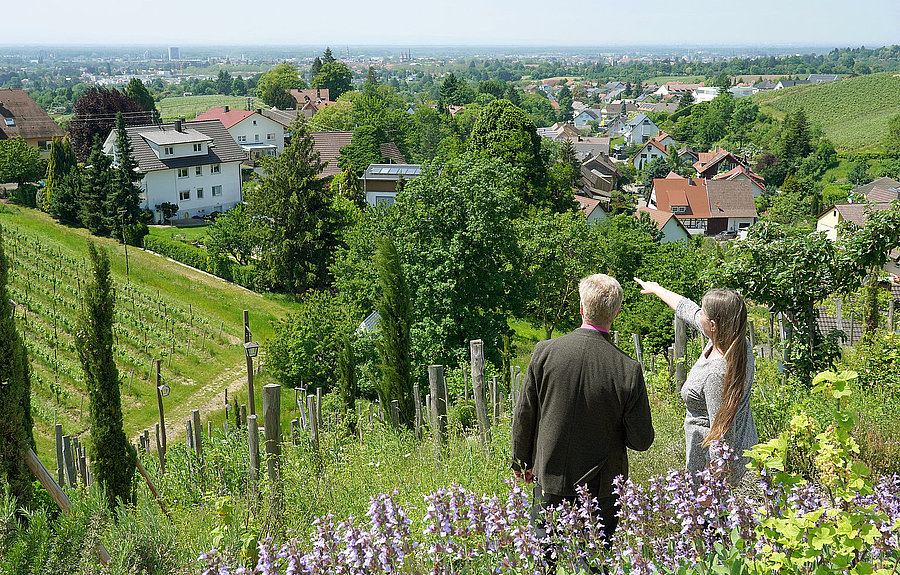 Mann und Frau besprechen sich. Sie stehen auf einem grünen Hügel und schauen auf eine Stadt.