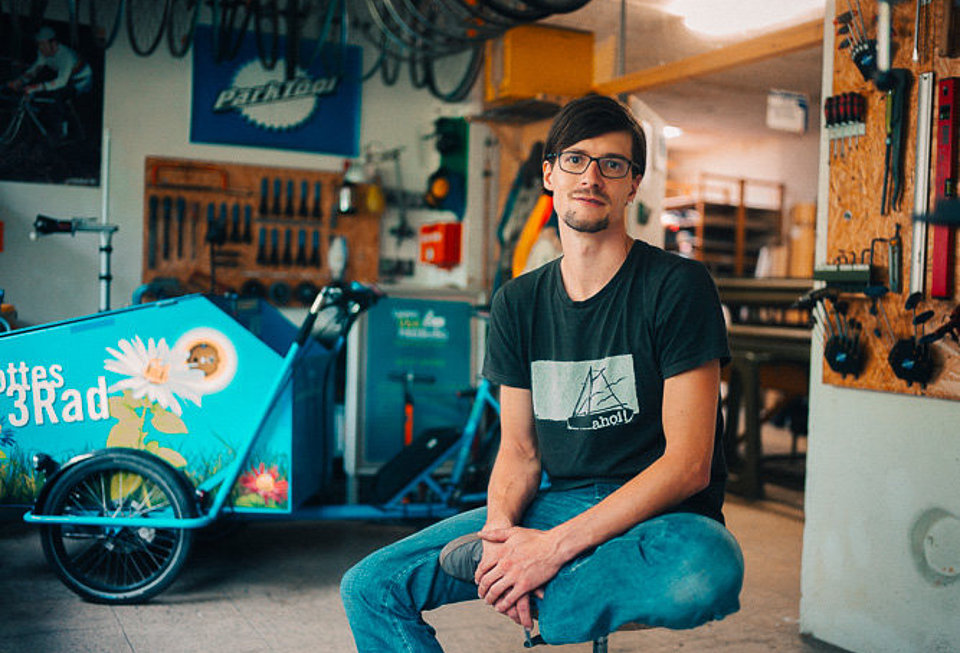 Junger Mann mit Brille sitzt in Werkstatt, im Hintergrund steht ein hellblaues Lastenrad