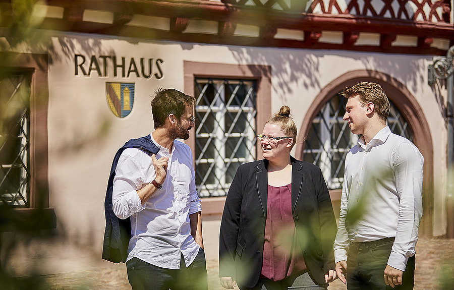 Zwei Männer und eine Frau stehen vor einem Rathaus und unterhalten sich