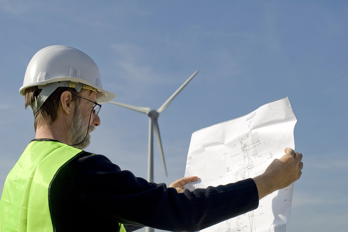 Mann mit Bauhelm steht in Windpark und schaut auf einen Plan