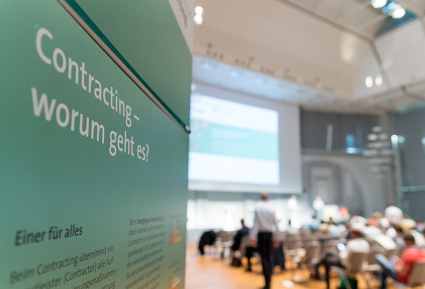 Aufsteller mit Aufschrift: Contracting-Kongress: Worum geht es? im Hintergrund sitzen Menschen auf Stühlen und schauen auf eine Leinwand