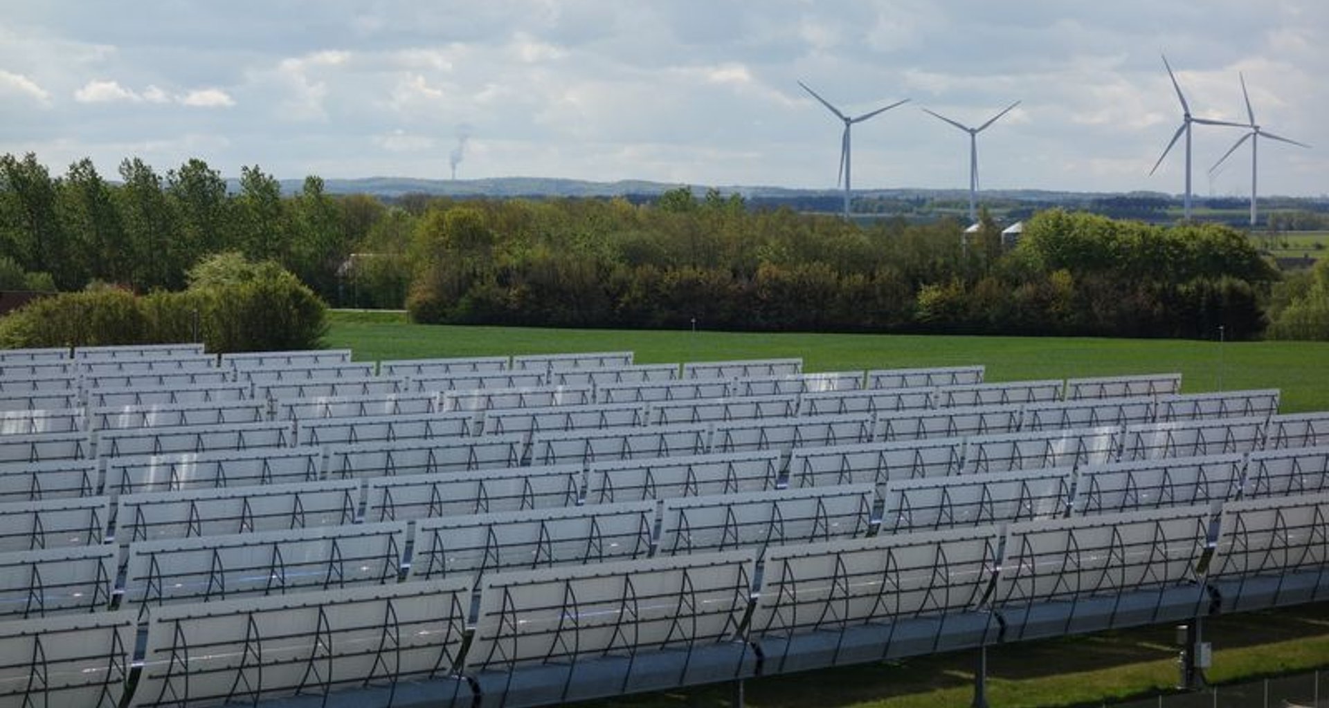 Foto eines Feldes auf dem im Vordergrund Solarpanelen und im Hintergrund Windräder zu sehen sind.