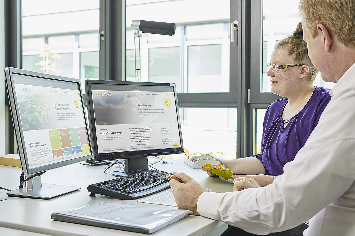 zwei personen sitzen am Schreibtisch vor zwei Bildschirmen