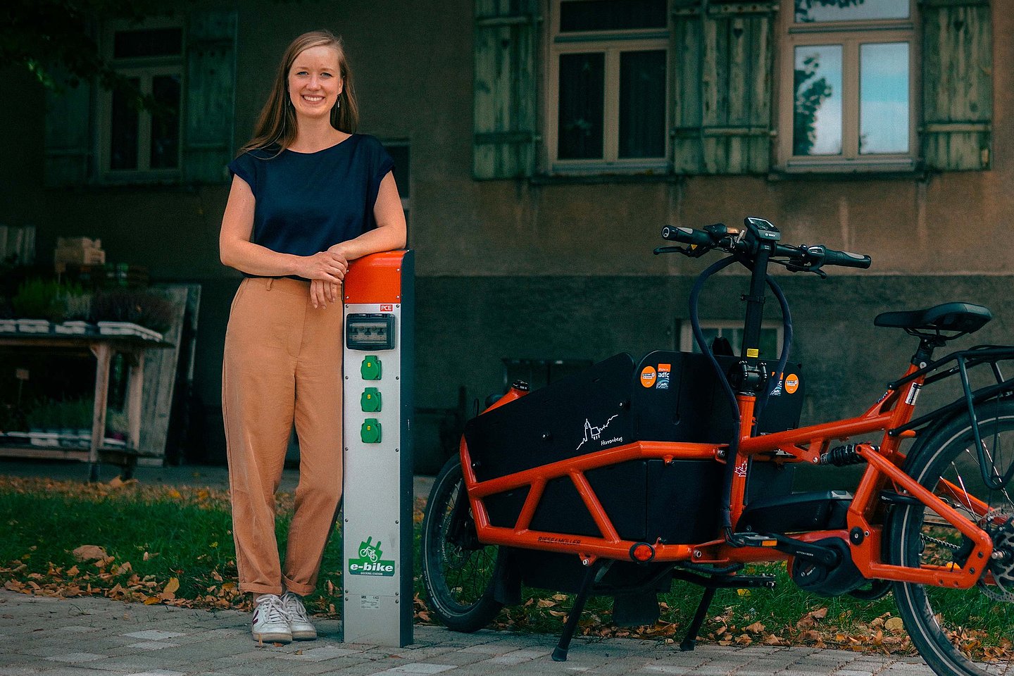 Das Bild zeigt eine Frau, die an einer Fahrradladesäule angelehnt ist. Neben ihr steht ein Lastenrad. 