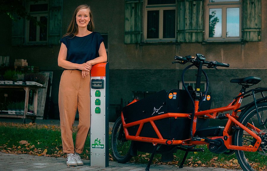 Das Bild zeigt eine Frau, die an einer Fahrradladesäule angelehnt ist. Neben ihr steht ein Lastenrad. 