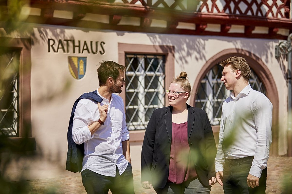 Drei Menschen beraten sich vor einem Rathaus.