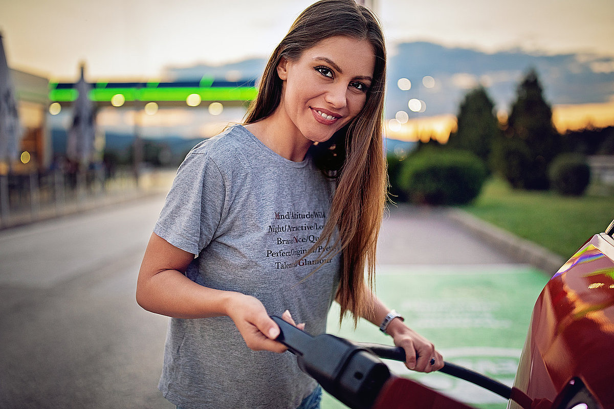 Eine junge Frau steht neben einem Auto und hält eine LAdestecker in der rechten Hand. Sie hat lange dunkle Haare und trägt ein graues T-Shirt. Foto: praetorianphoto/iStock 