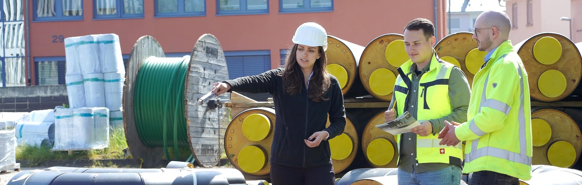 Drei Personen stehen auf einer Baustelle vor gestapelten, isolierten Rohren für ein Wärmenetz. Eine Frau mit Bauhelm zeigt auf ein Bauteil, während zwei Kollegen in gelber Arbeitsplanung zuhören.