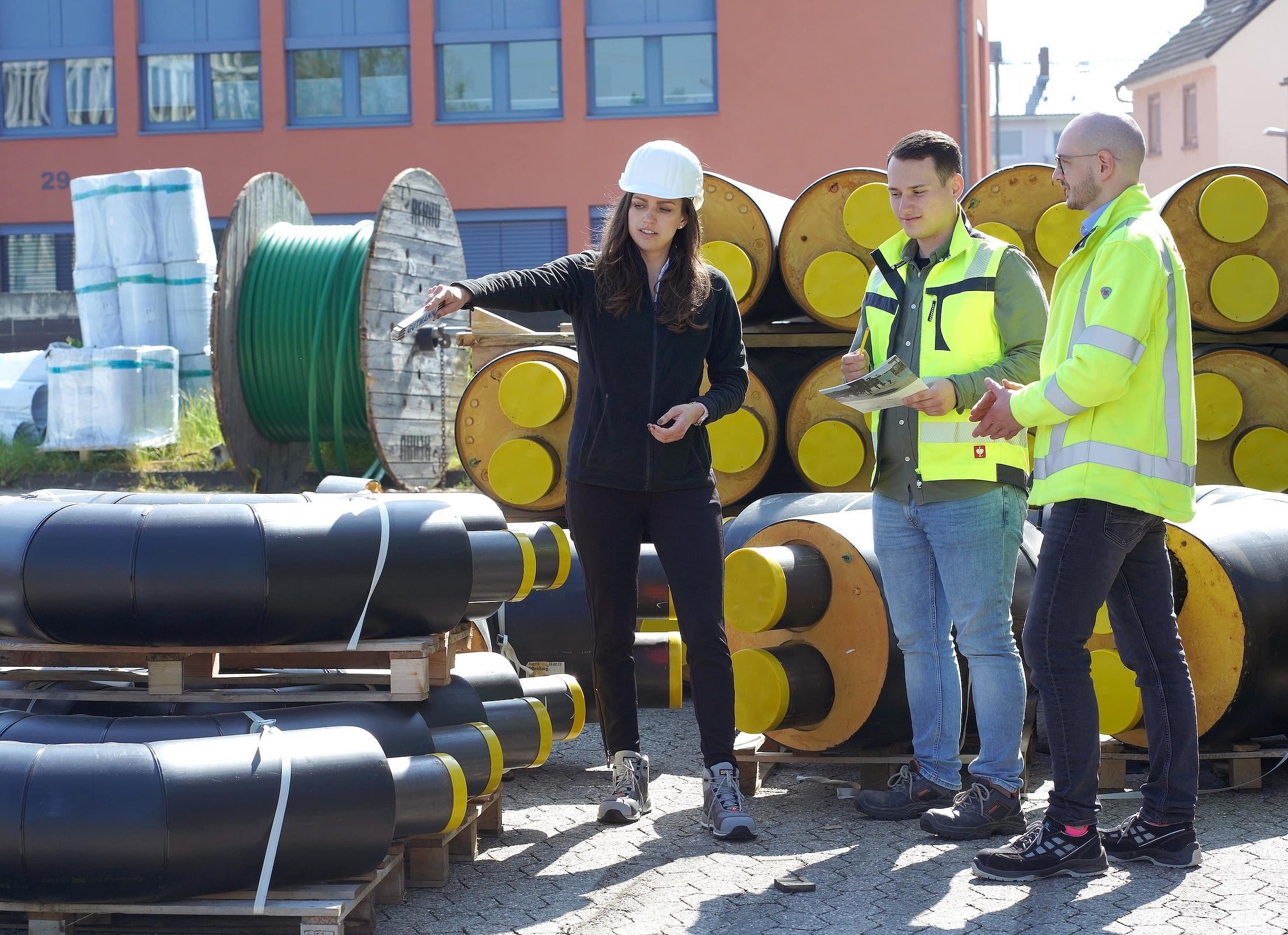Drei Personen stehen auf einer Baustelle vor gestapelten, isolierten Rohren für ein Wärmenetz. Eine Frau mit Bauhelm zeigt auf ein Bauteil, während zwei Kollegen in gelber Arbeitsplanung zuhören.