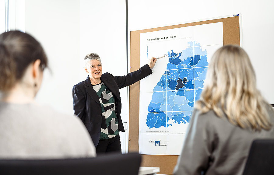 Eine Frau steht vor einem Schaublid und erklärt dieses einer Gruppe