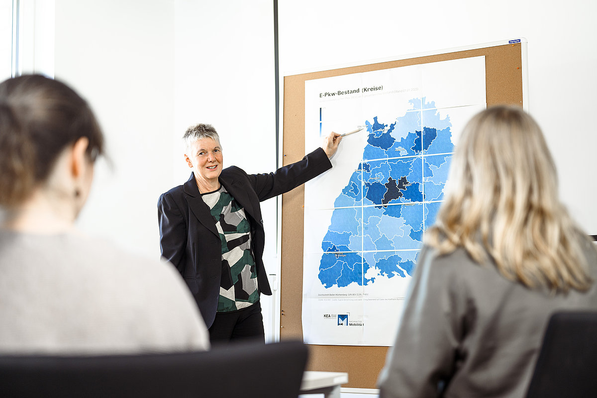 Eine Frau steht vor einem Schaublid und erklärt dieses einer Gruppe