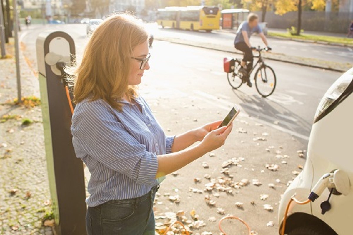 Frau mit Smartphone beim Laden des Autos vor Fahrradfahrer und Bus im Hintergrund