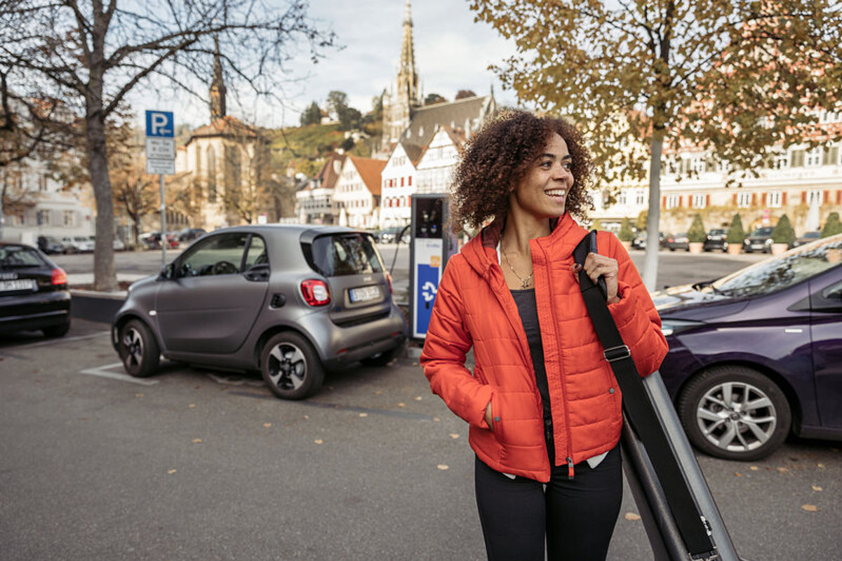 Eine jugne Frau in der Stadt, hinter ihr sind zwei Autos an eine Ladesäule angeschlossen.
