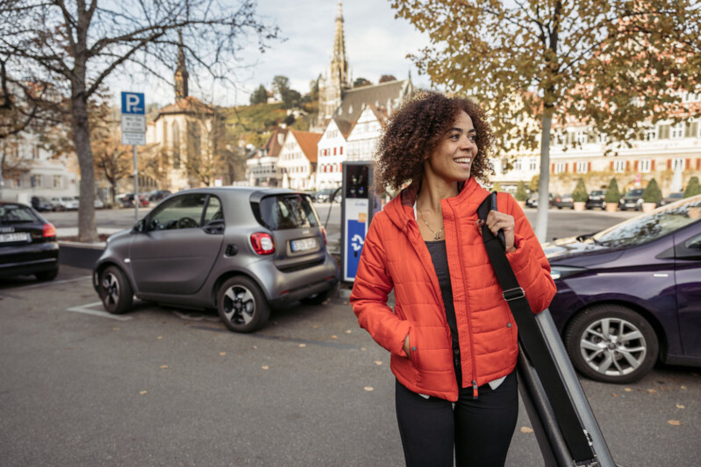 Eine jugne Frau in der Stadt, hinter ihr sind zwei Autos an eine Ladesäule angeschlossen.