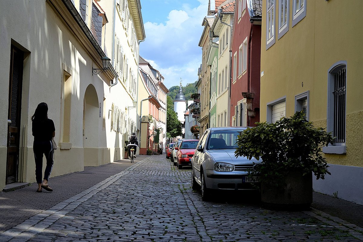 Blick auf eine Gasse in Heidelberg mit Fußgängerin, Radfahrer und parkenden Autos