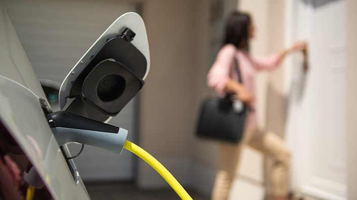 E-Auto mit Ladekabel, Frau im Hintergrund vor Haustür