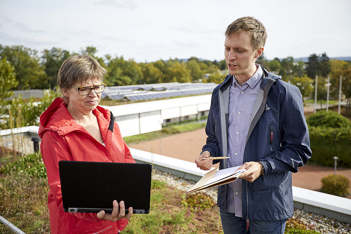 Frau und Mann stehen auf einem Schuldach mit Photovoltaik und schauen in einen Laptop