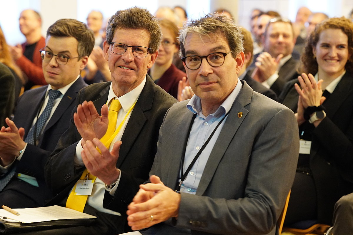 Drei Männer im Vordergrund vor größerem Publikum klatschen