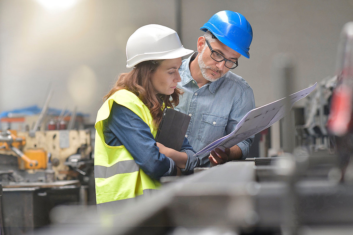 Frau und Mann mit Bauhelm besprechen sich in einem Industriebetrieb über einen Plan gebeugt