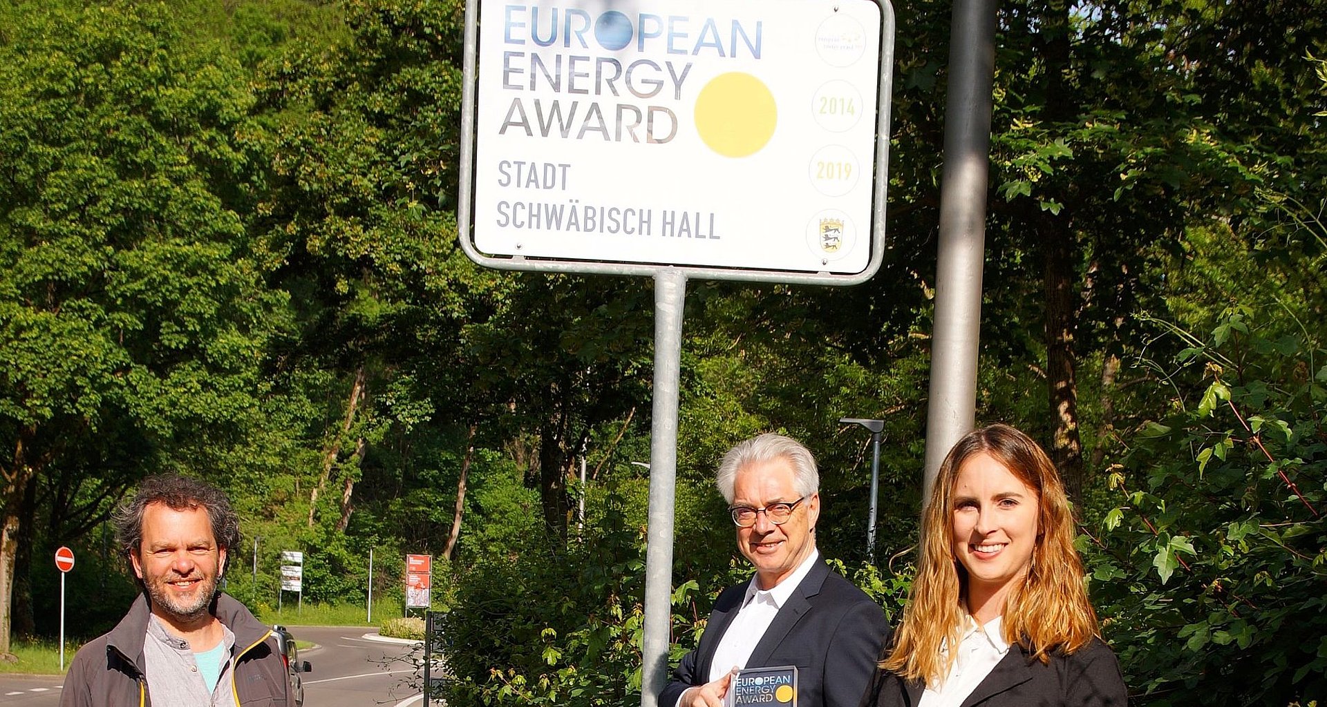 zwei Männer und eine Frau stehen vor dem Straßenschild der Goldauszeichnung "European Energie Award"