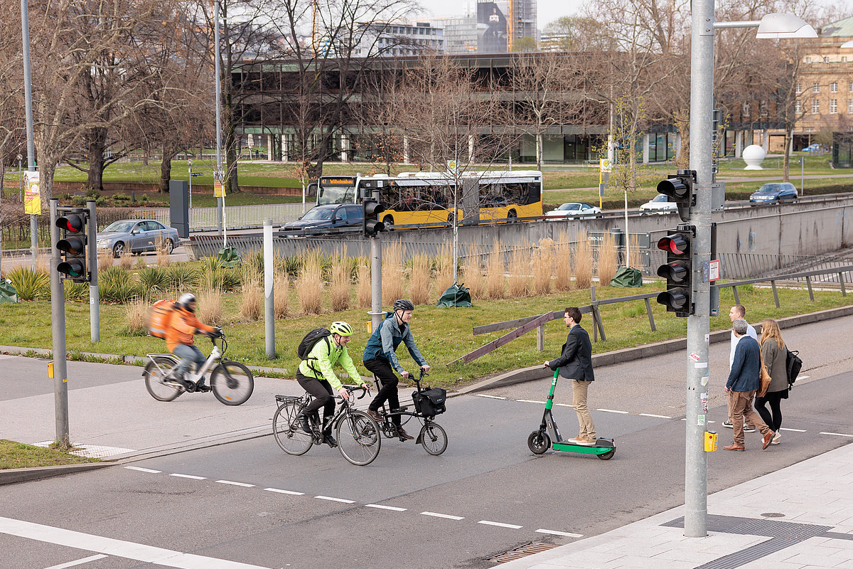 Menschen queren Ampel zu Fuß, per Rad und Roller vor dem Stuttgarter Landtag, Bus und Pkw im Hintergrund