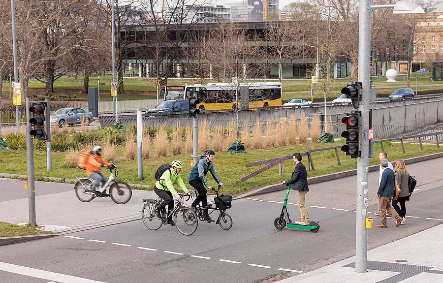 Menschen queren Ampel zu Fuß, per Rad und Roller vor dem Stuttgarter Landtag, Bus und Pkw im Hintergrund