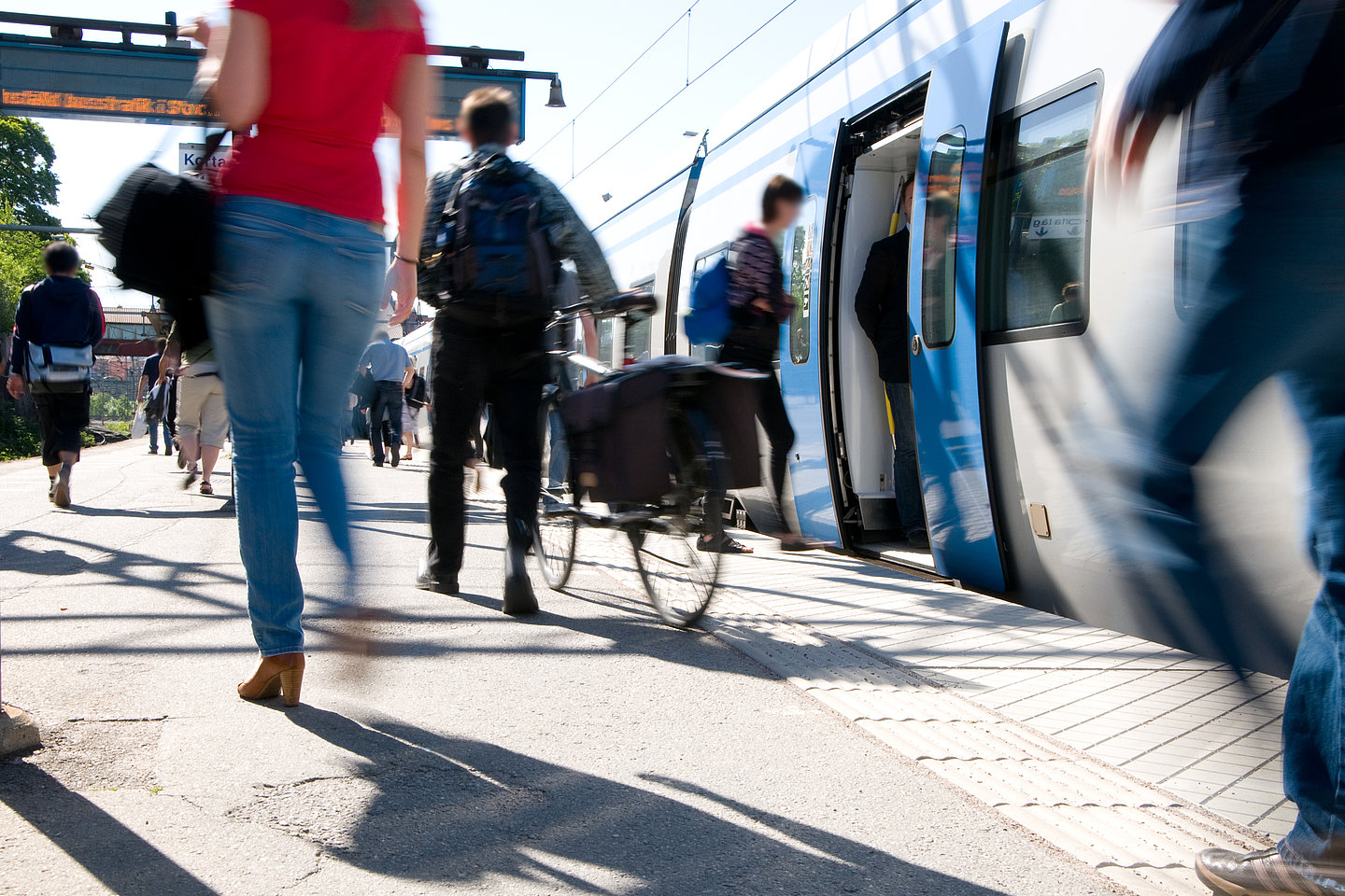 Viele Menschen sind unterwegs bei einem Bahnhof. Einigen steigen in einen Zug im Hintergrund ein.