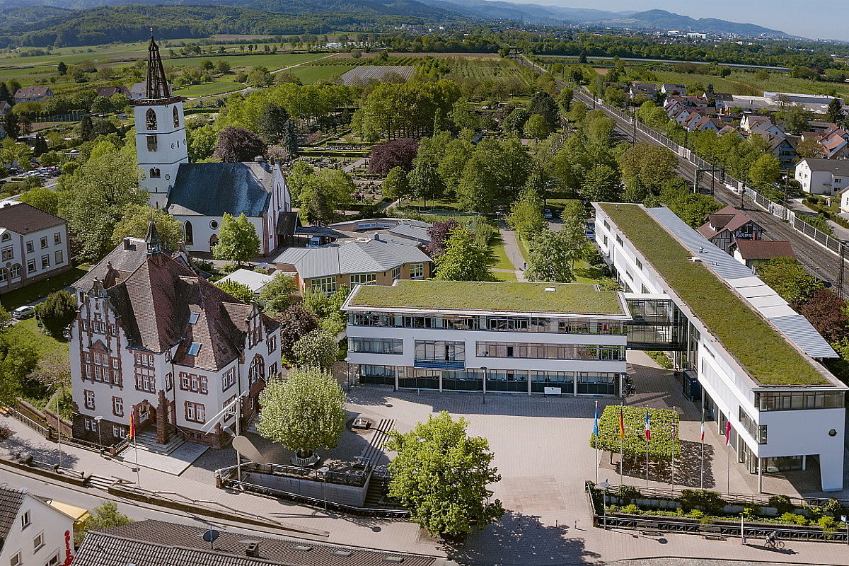 Luftbild einer kleinen Gemeinde mit Rathaus, Kirche und begrünten Dächern. Im Hintergrund der Schwarzwald.