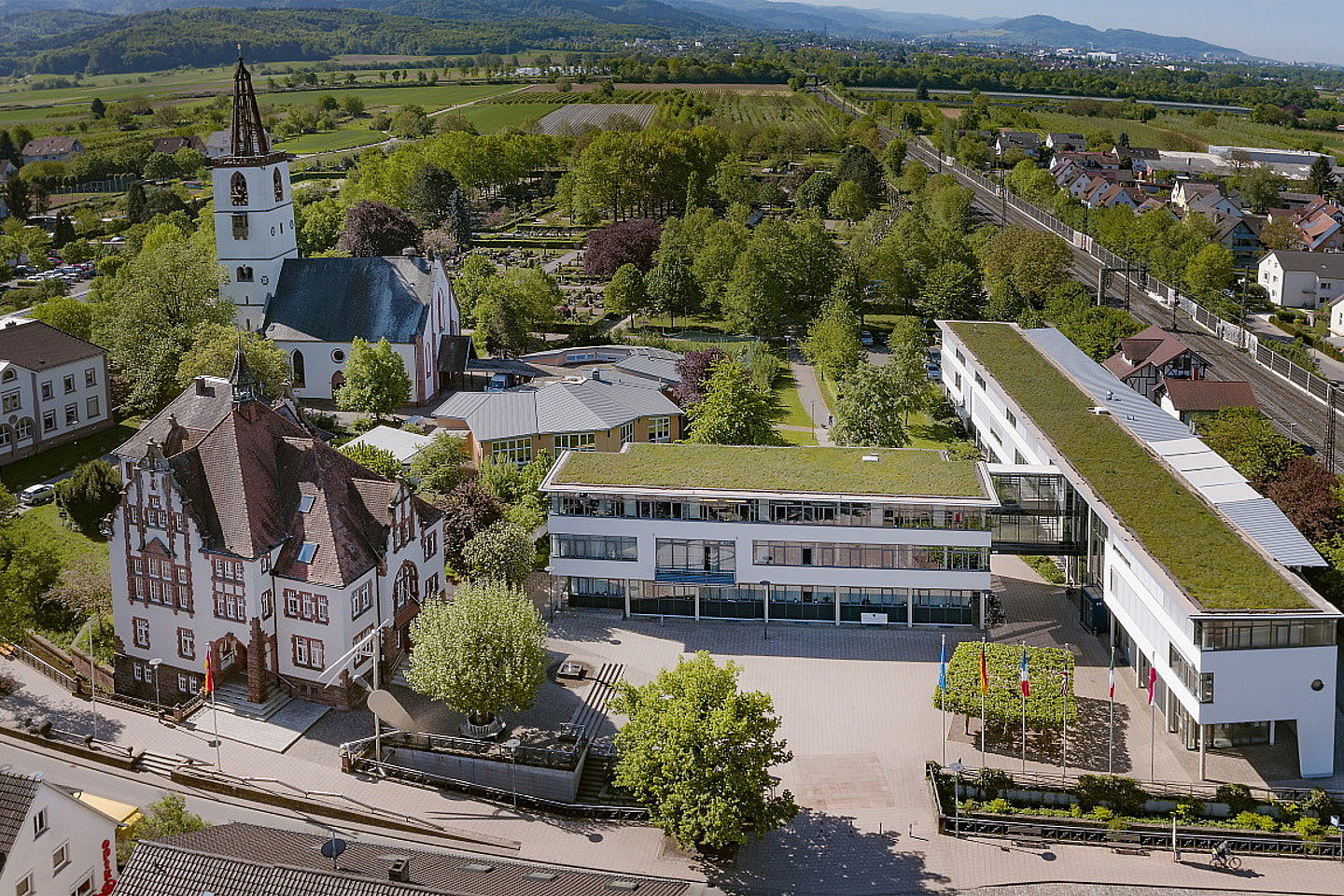 Luftbild einer kleinen Gemeinde mit Rathaus, Kirche und begrünten Dächern. Im Hintergrund der Schwarzwald.