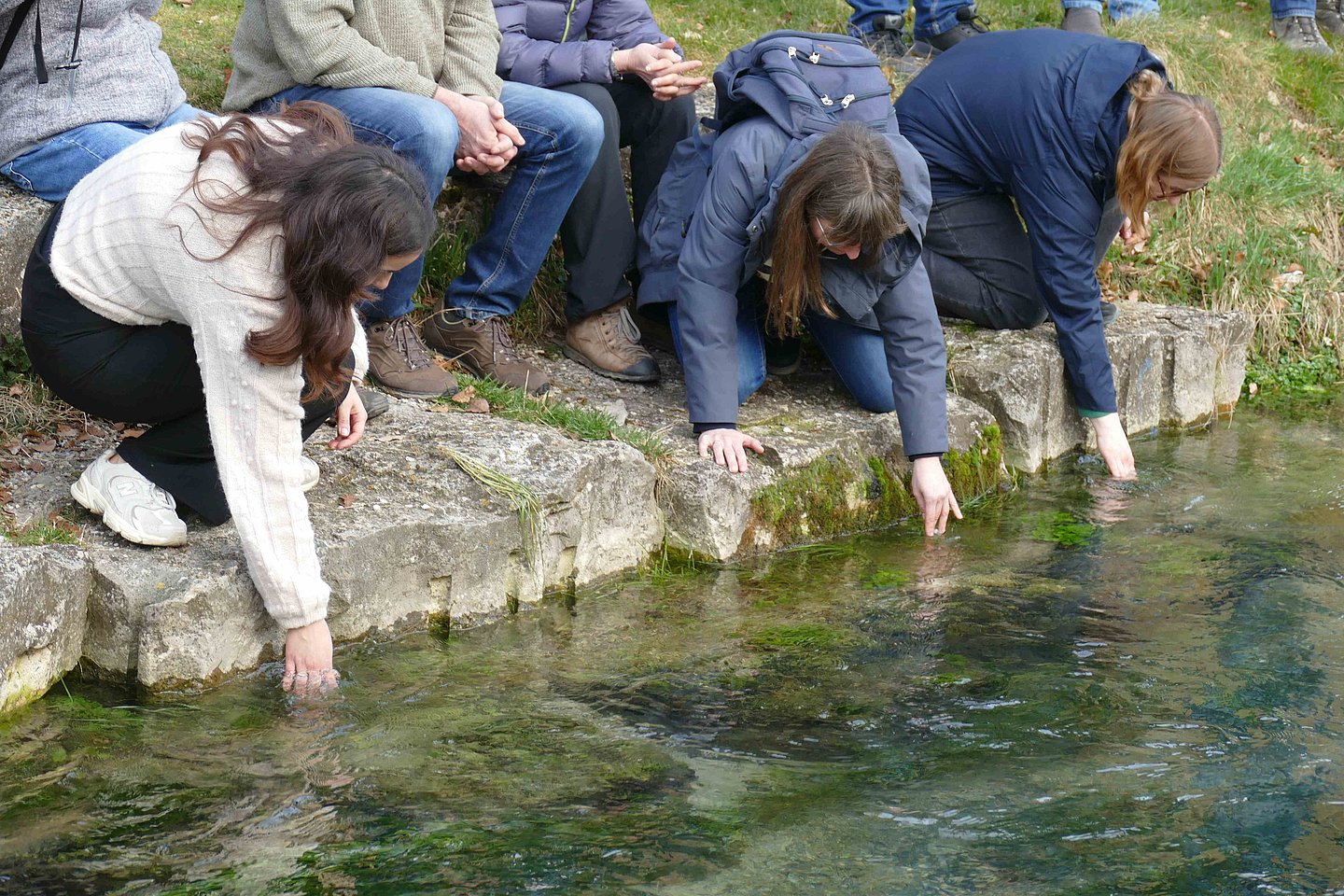 Drei Personen hocken am Gewässerrand und fassen ins Wasser